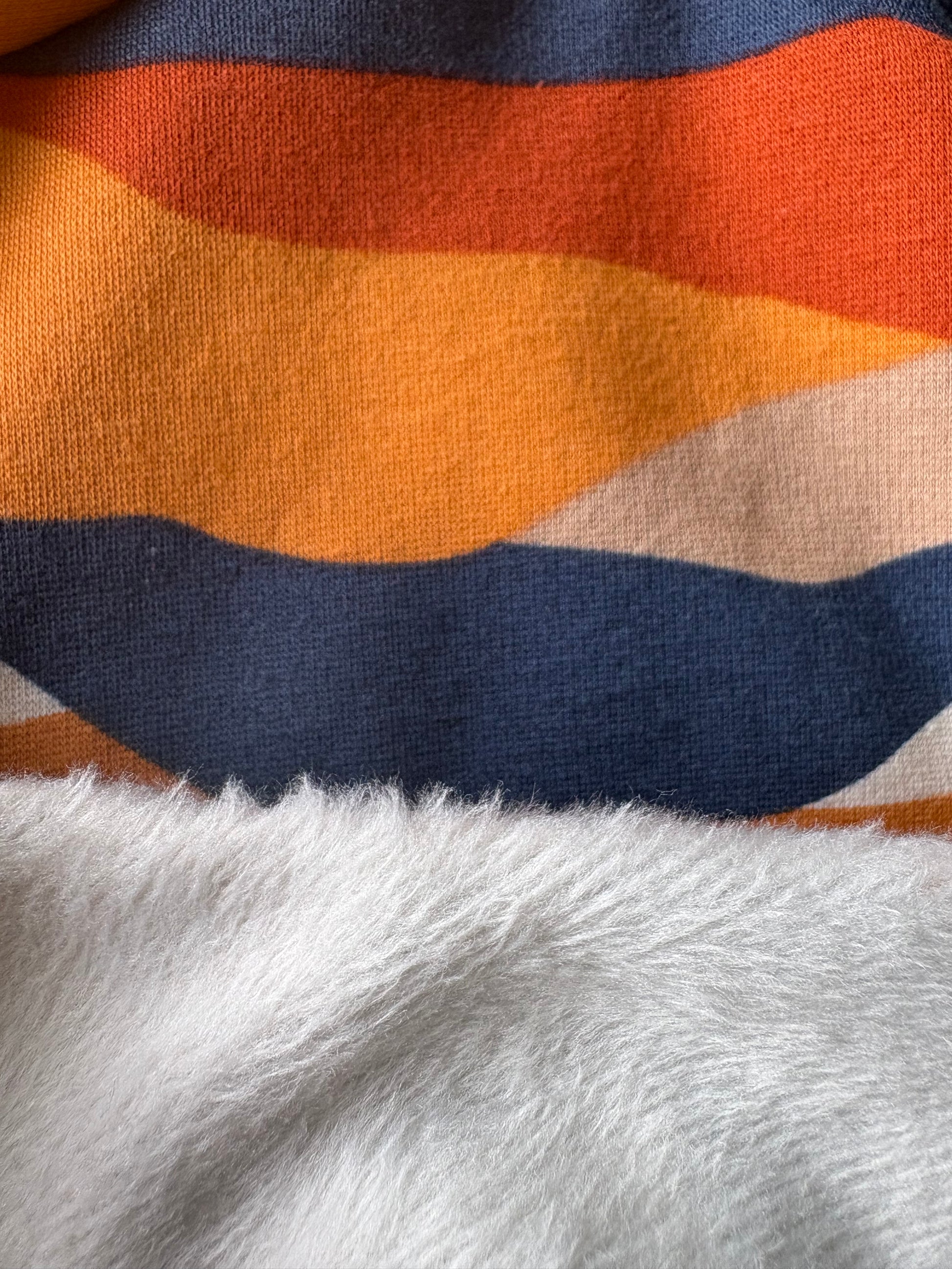 Close-up of a knit fabric with wavy horizontal stripes in orange, navy blue, beige, and white, with a soft white faux-fur texture layered along the bottom edge.